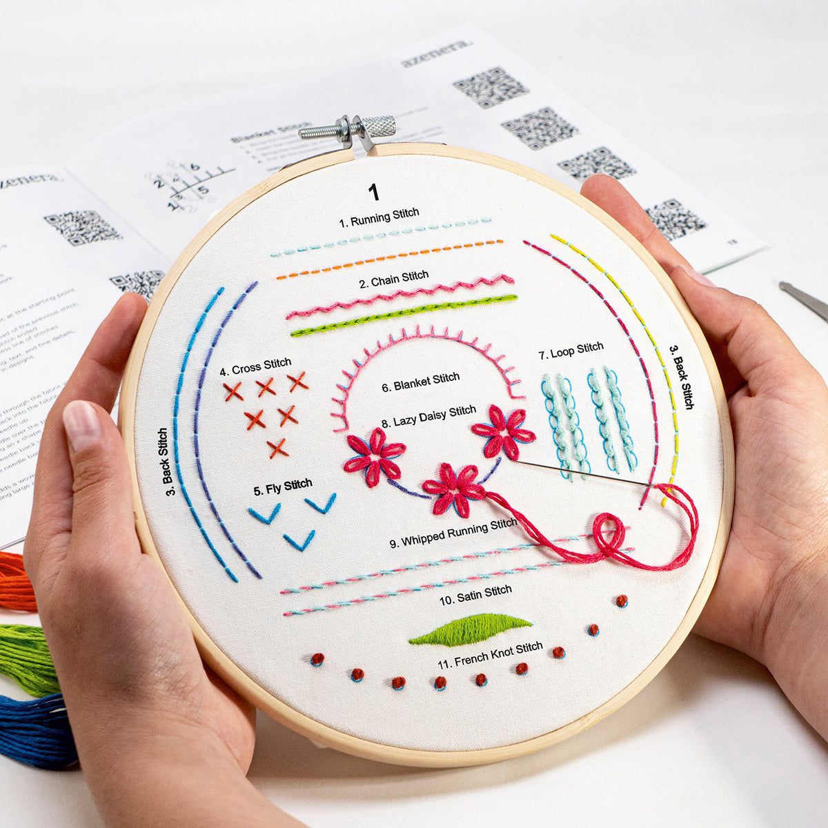 A pair of hands holds a wooden embroidery hoop displaying a labeled stitch guide with colorful sample stitches on white fabric. Instruction sheets are visible in the background on a white surface.