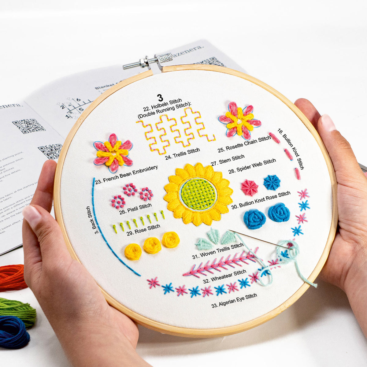 A person holds a round embroidery hoop containing a white fabric sample with colorful embroidered stitch patterns and printed stitch names. An open instruction booklet is visible behind the hoop on a white surface.