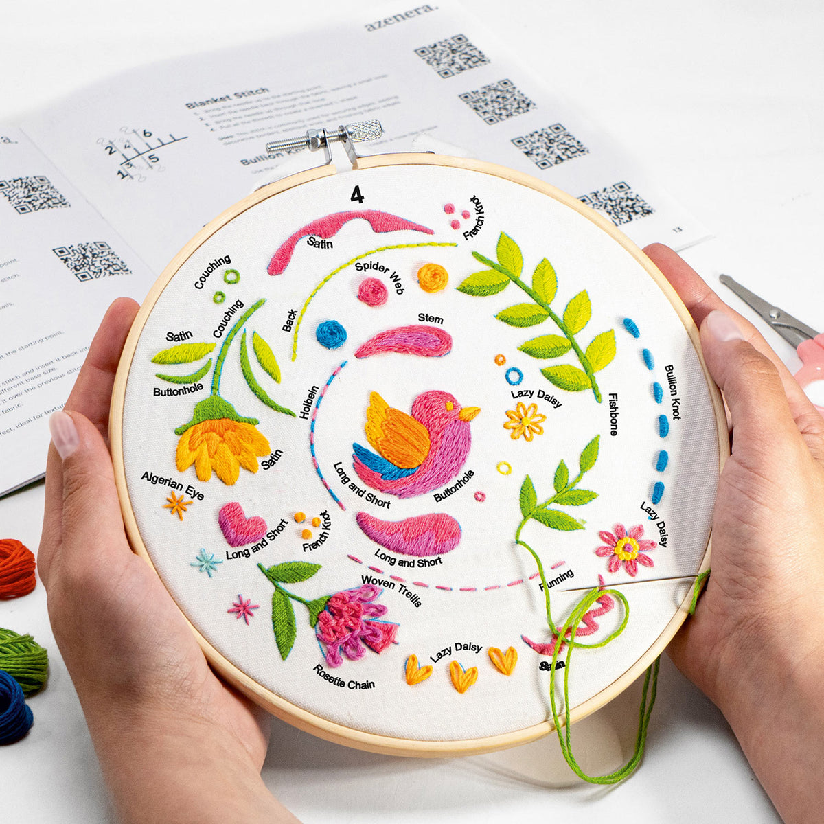 A person holds a wooden embroidery hoop containing a colorful embroidered bird and floral design on white fabric. An instruction booklet and scissors are visible in the background on a white surface.